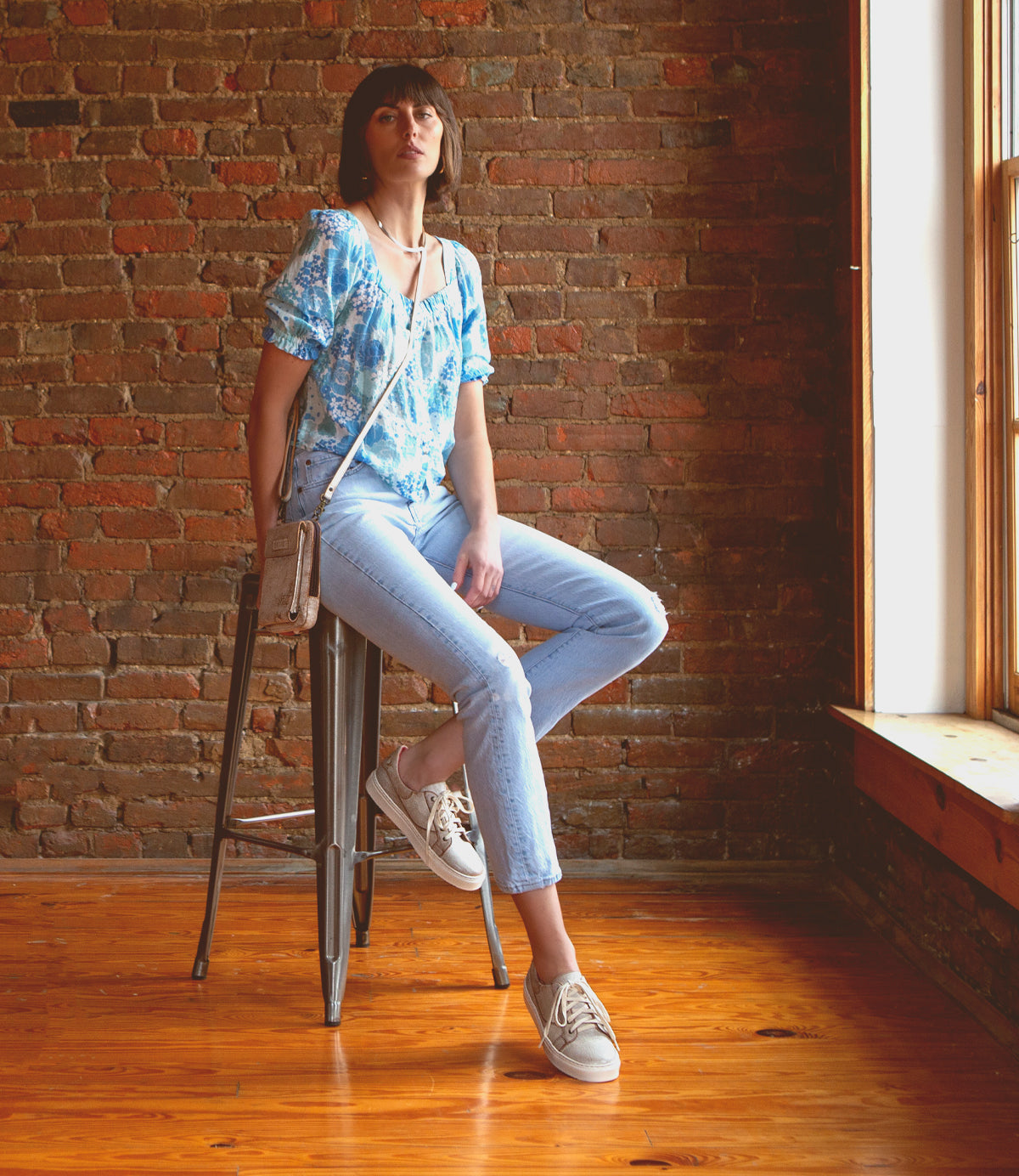 A woman in a blue floral top and light jeans sits on a stool by a brick wall and window, wearing the Bed|Stü Azeli leather sneakers with a durable rubber outsole for all-day comfort and carrying a crossbody bag.