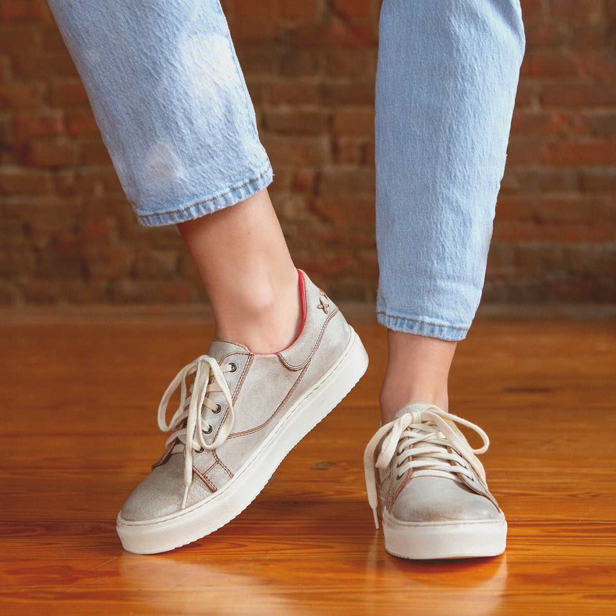 A person wearing the Bed|Stü Azeli light gray canvas sneakers with white laces and light blue jeans stands on a wooden floor against a brick wall background. The Azeli sneakers feature a durable rubber outsole for lasting wear and all-day comfort.
