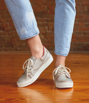A person wearing the Bed|Stü Azeli light gray canvas sneakers with white laces and light blue jeans stands on a wooden floor against a brick wall background. The Azeli sneakers feature a durable rubber outsole for lasting wear and all-day comfort.