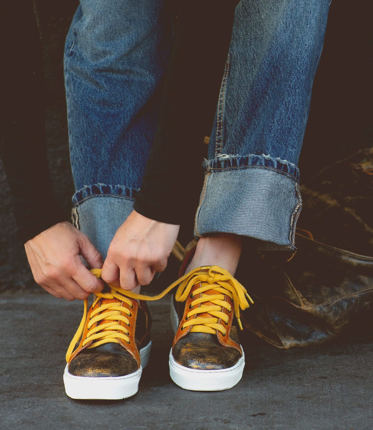 A person tying yellow shoelaces on the Bed|Stü Azeli metallic leather sneakers, featuring a durable rubber outsole and styled with blue cuffed jeans—perfect for all-day comfort.