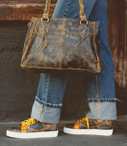 A person wearing rolled-up blue jeans and Bed|Stü’s Azeli brown leather sneakers—with durable rubber outsoles for all-day comfort and distinctive yellow laces—carries a distressed brown leather handbag while standing on a gray sidewalk.