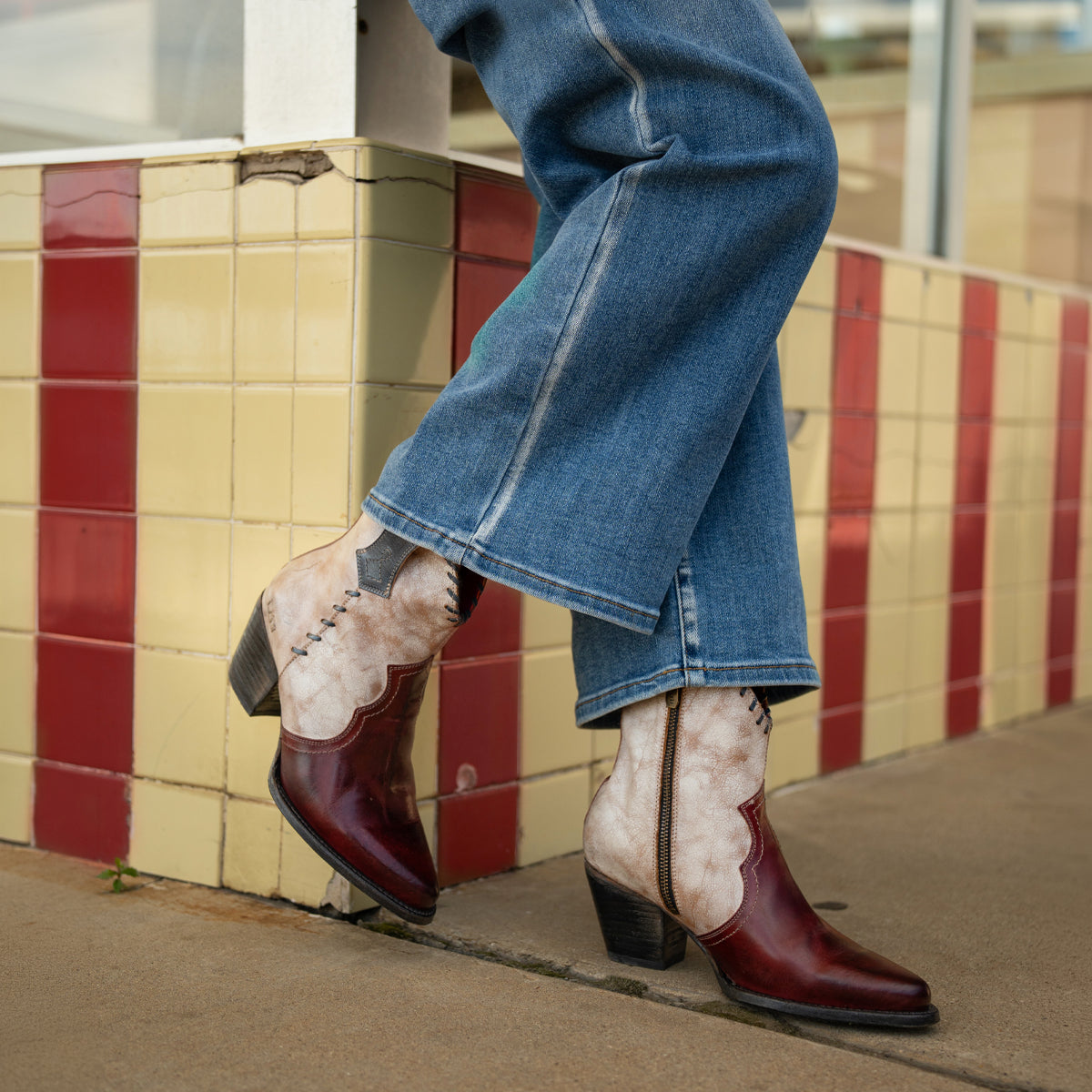 A person wearing flared jeans and the Bed|Stü Baila II two-tone red and tan Western-inspired, pointed-toe boots stands on a sidewalk in front of a tiled wall.