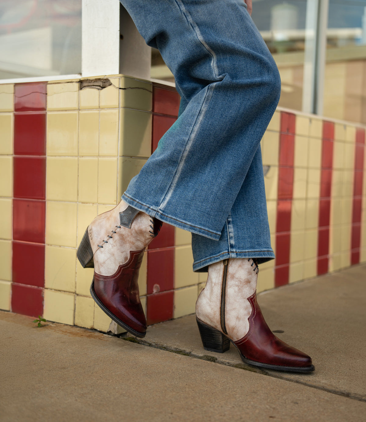 A person wearing flared jeans and the Bed|Stü Baila II two-tone red and tan Western-inspired, pointed-toe boots stands on a sidewalk in front of a tiled wall.