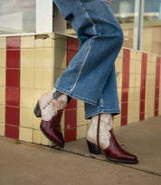 A person wearing flared jeans and the Bed|Stü Baila II two-tone red and tan Western-inspired, pointed-toe boots stands on a sidewalk in front of a tiled wall.