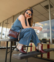 A woman in a blue denim jumpsuit and red boots sits on a picnic table, holding a drink. She accessorizes with white sunglasses, a blue headband, and carries a brown purse. Her Bed|Stü Baila II Western-inspired boots make a bold statement to complete the look.