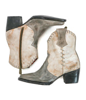 A pair of Bed|Stü Baila II distressed tan and gray cowboy boots, featuring pointed toes, block heels, visible stitching, and side zippers, showcase artisanal construction and Western-inspired design, set against a white background.