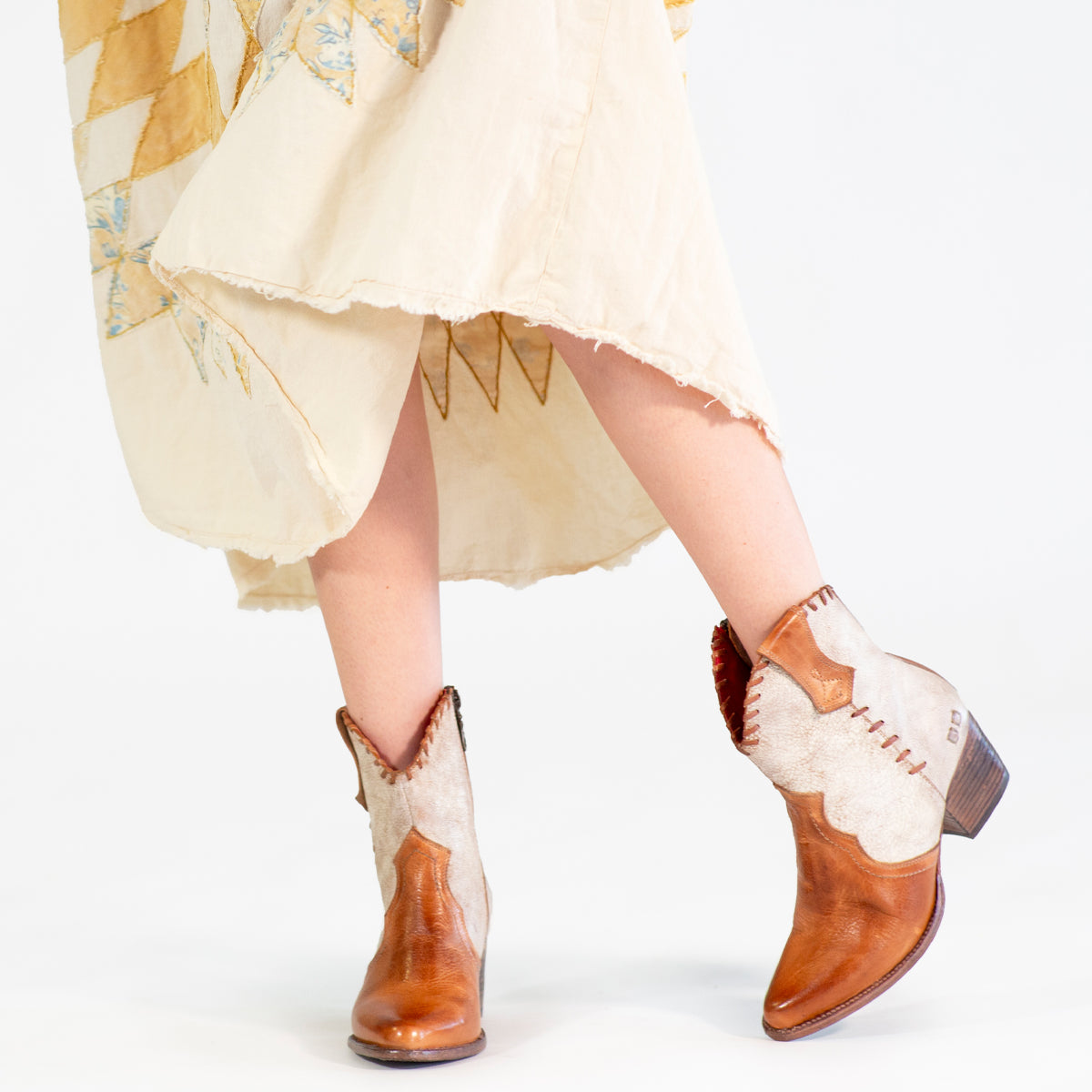 A person wearing a cream patchwork dress and the Bed|Stü Baila II light brown Western-inspired boots with a pointed toe, standing with legs crossed against a white background.