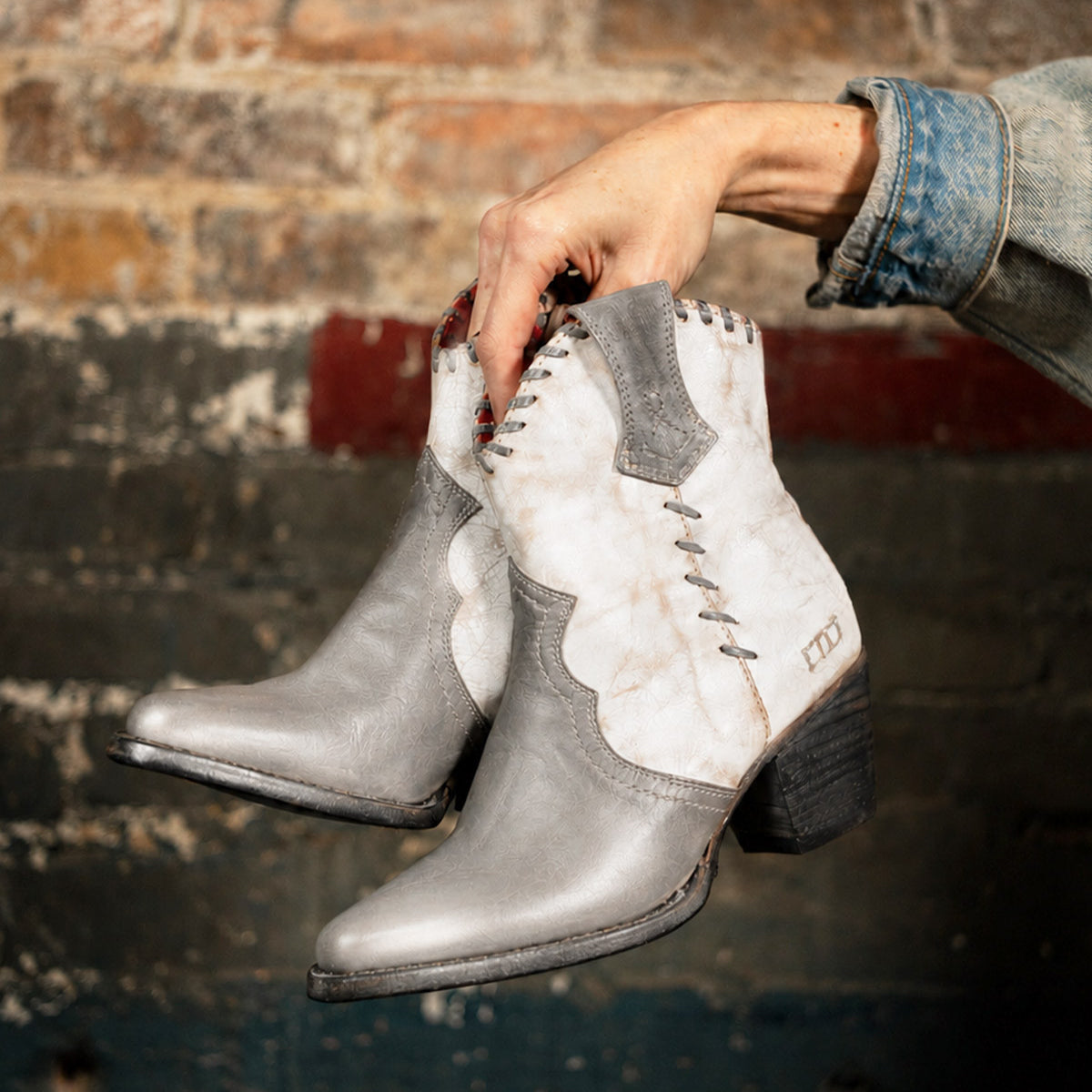 A hand holds the Bed|Stü Baila II boots—gray and white leather, Western-inspired with intricate stitching—set against a worn brick and painted wall background.