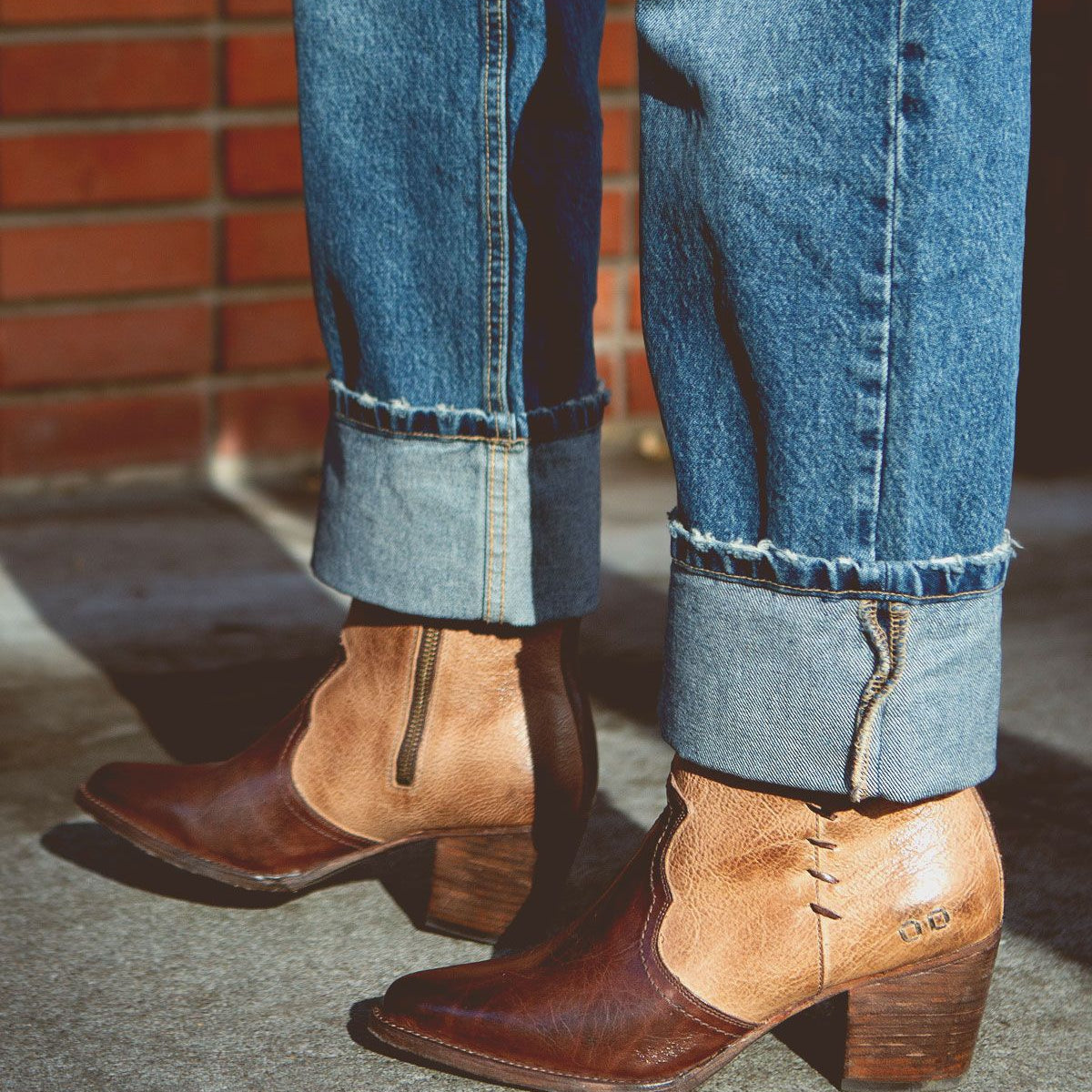 Wearing cuffed blue jeans and the Bed|Stü Baila II two-tone brown leather ankle boots with block heels—artfully crafted and subtly Western-inspired—the person stands on a concrete surface beside a brick wall.