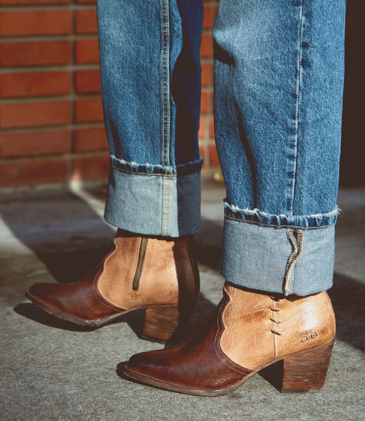 Wearing cuffed blue jeans and the Bed|Stü Baila II two-tone brown leather ankle boots with block heels—artfully crafted and subtly Western-inspired—the person stands on a concrete surface beside a brick wall.