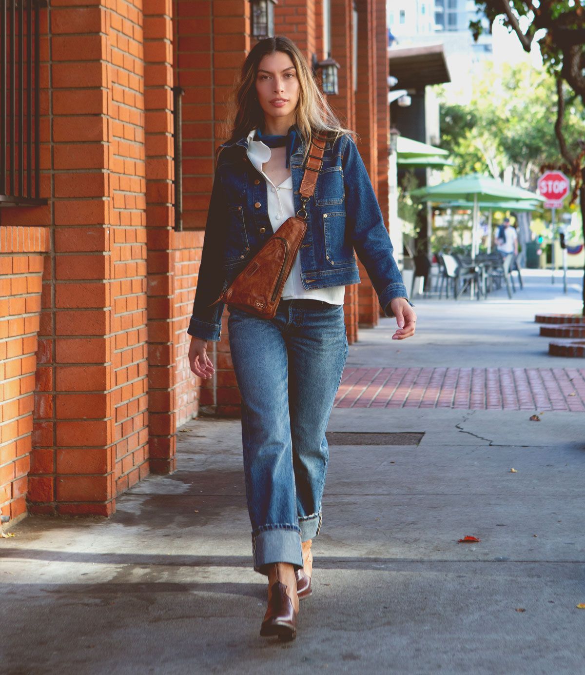 A woman wearing a denim jacket, white shirt, blue jeans, and Bed|Stü Baila II Western-inspired boots with a pointed toe walks on a sidewalk next to a brick building, carrying a brown crossbody bag.
