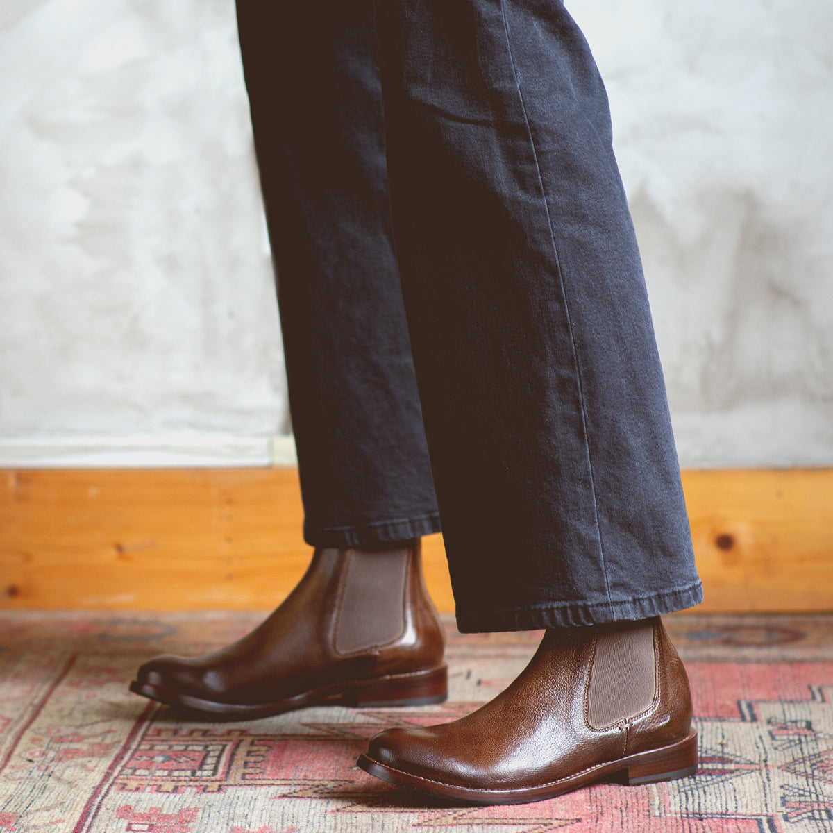 A person wearing dark pants and Bed|Stü Barfly brown Italian leather Chelsea boots stands on a patterned rug against a textured wall.