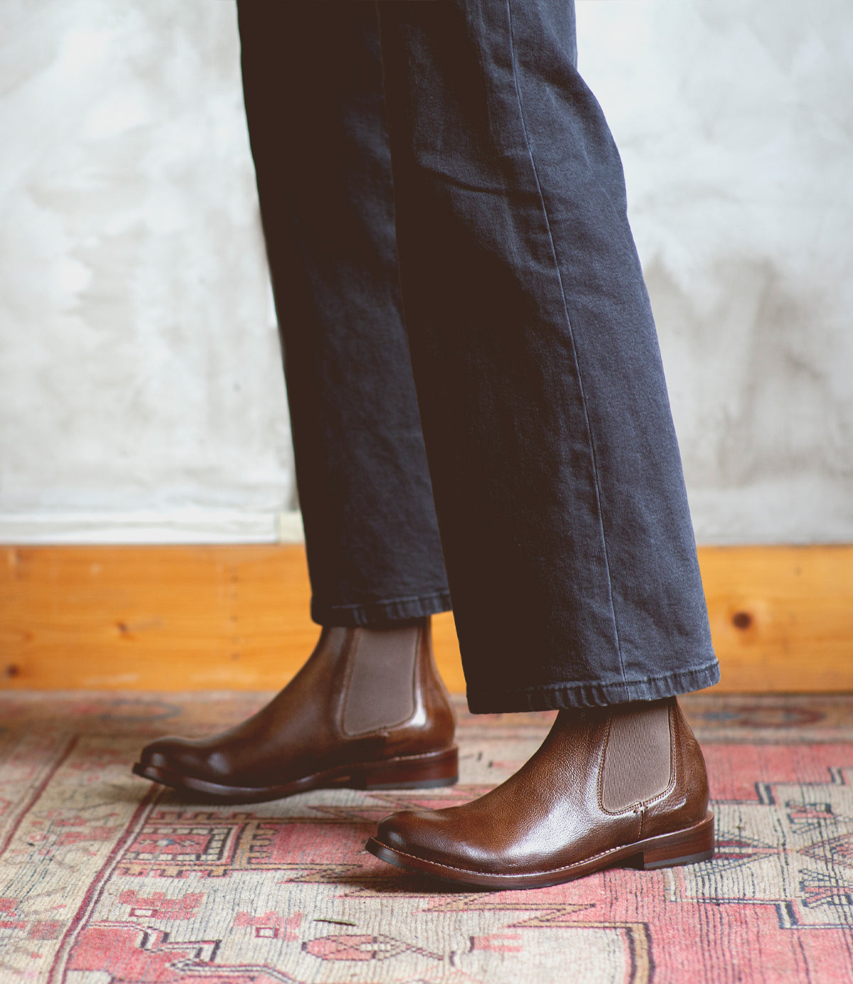 A person wearing dark pants and Bed|Stü Barfly brown Italian leather Chelsea boots stands on a patterned rug against a textured wall.