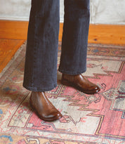 A person wearing dark pants and Bed|Stü Barfly brown Italian leather Chelsea boots stands on a patterned rug featuring red, pink, and beige tones.