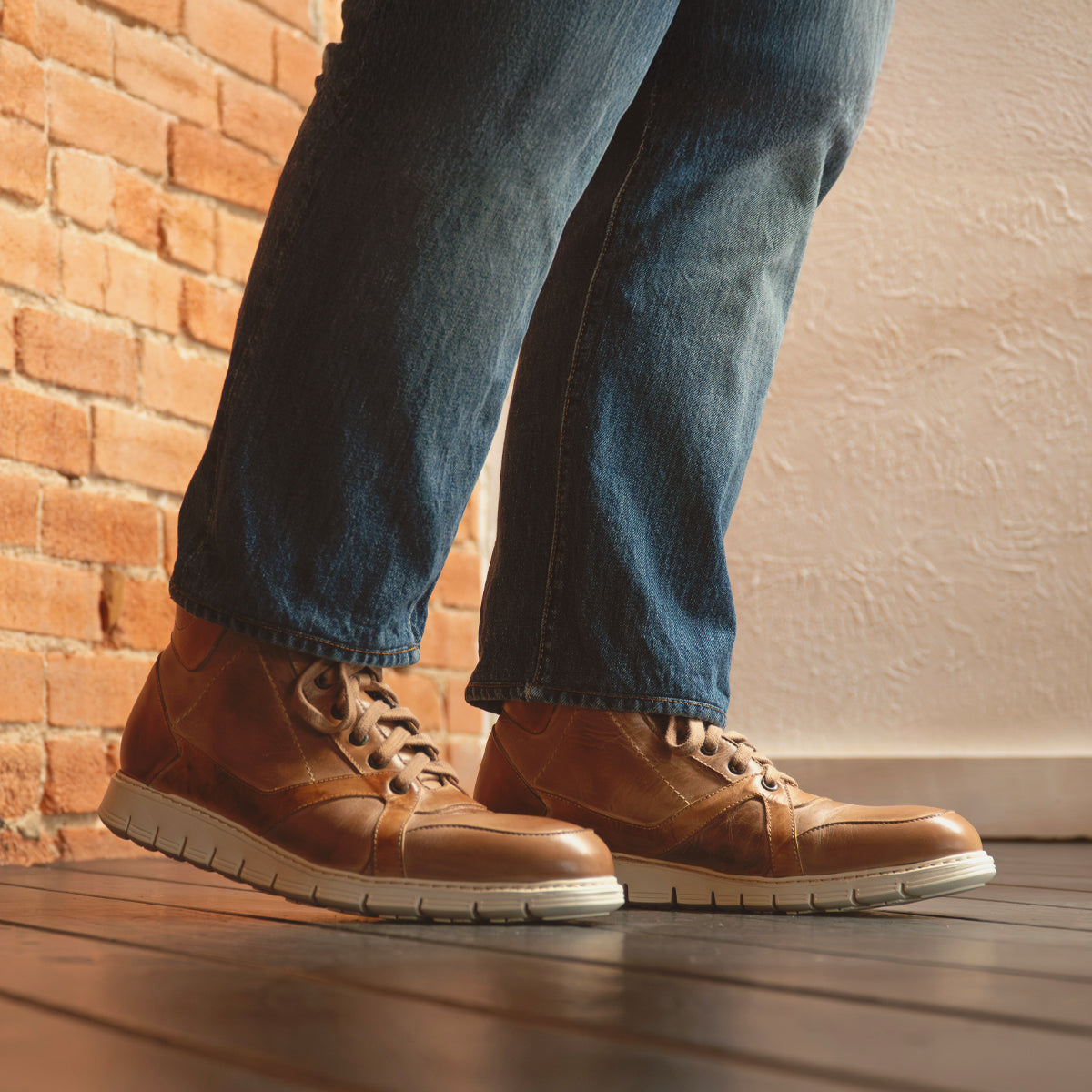 A person wearing blue jeans and Bed|Stü Barton brown two-tone leather high-top sneakers with a lightweight sole stands on a wooden floor next to a brick wall.
