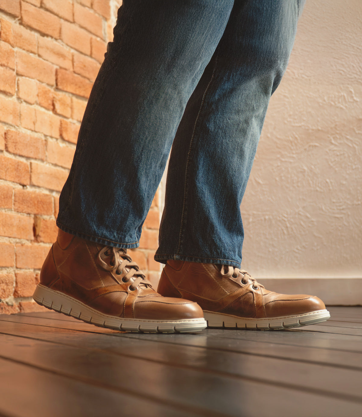 A person wearing blue jeans and Bed|Stü Barton brown two-tone leather high-top sneakers with a lightweight sole stands on a wooden floor next to a brick wall.
