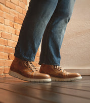 A person wearing blue jeans and Bed|Stü Barton brown two-tone leather high-top sneakers with a lightweight sole stands on a wooden floor next to a brick wall.