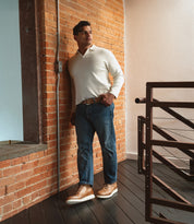 A man stands indoors by a brick wall, wearing a white sweater and blue jeans, with his hands in his pockets, looking to the side. He completes the look with Bed|Stü's Barton two-tone leather high-top sneakers featuring a lightweight sole.