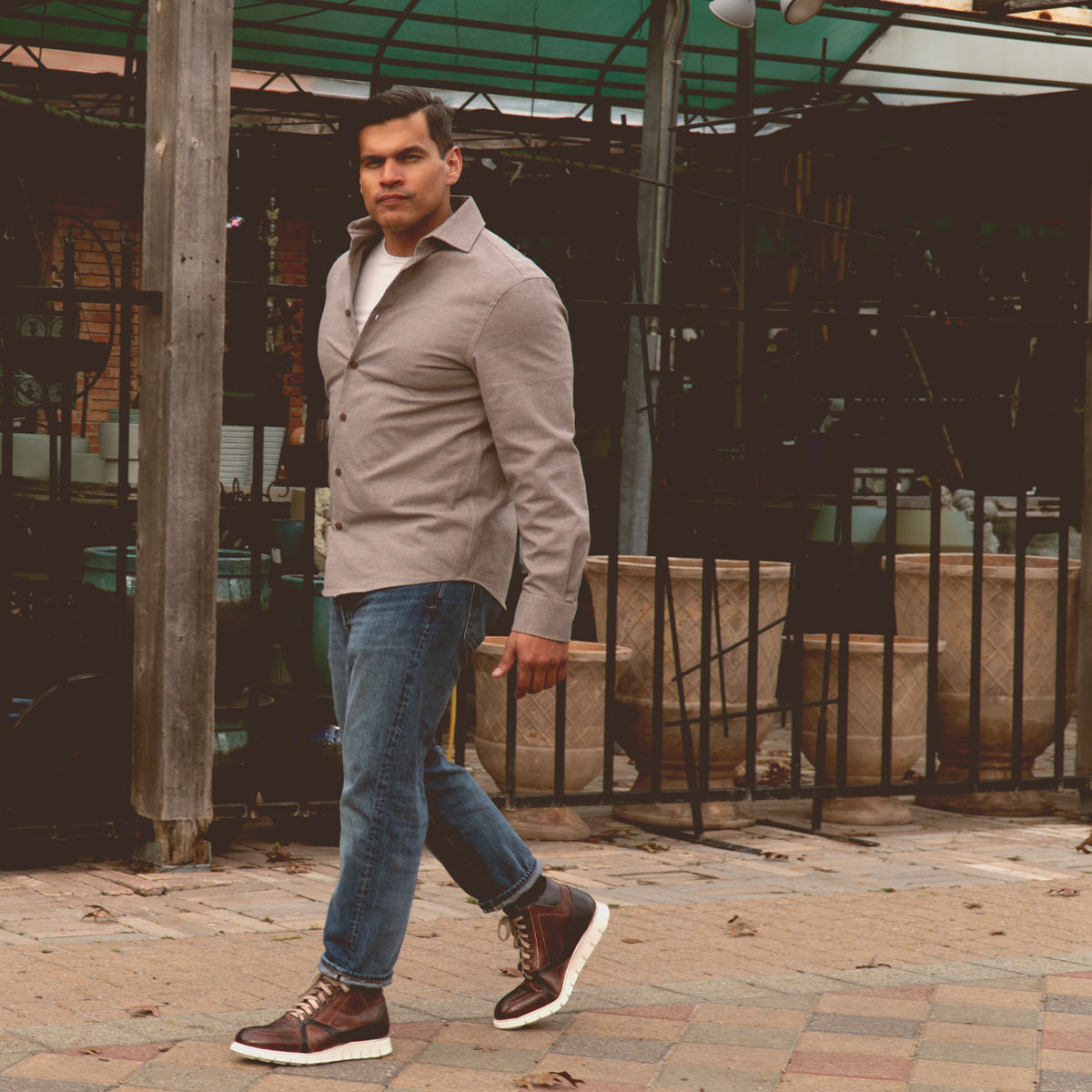 A man in a button-up shirt, jeans, and Bed|Stü Barton boots with a lightweight sole walks outdoors on a tiled surface near potted plants and wooden structures.