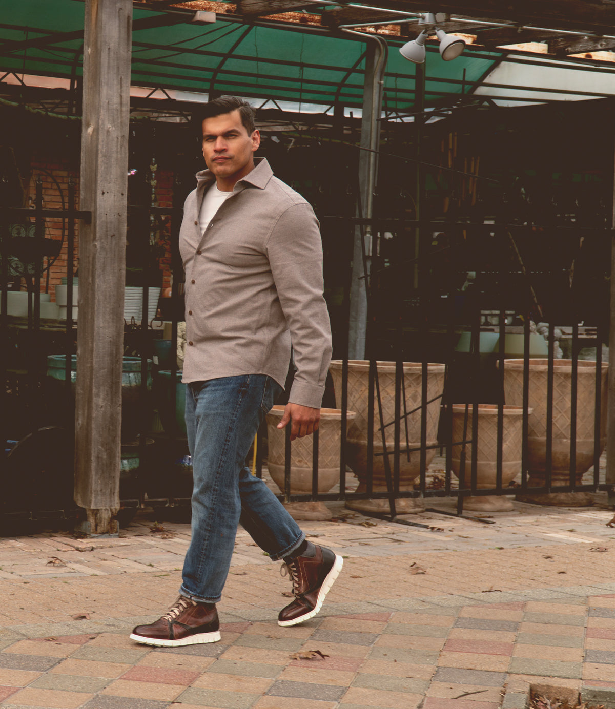 A man in a button-up shirt, jeans, and Bed|Stü Barton boots with a lightweight sole walks outdoors on a tiled surface near potted plants and wooden structures.