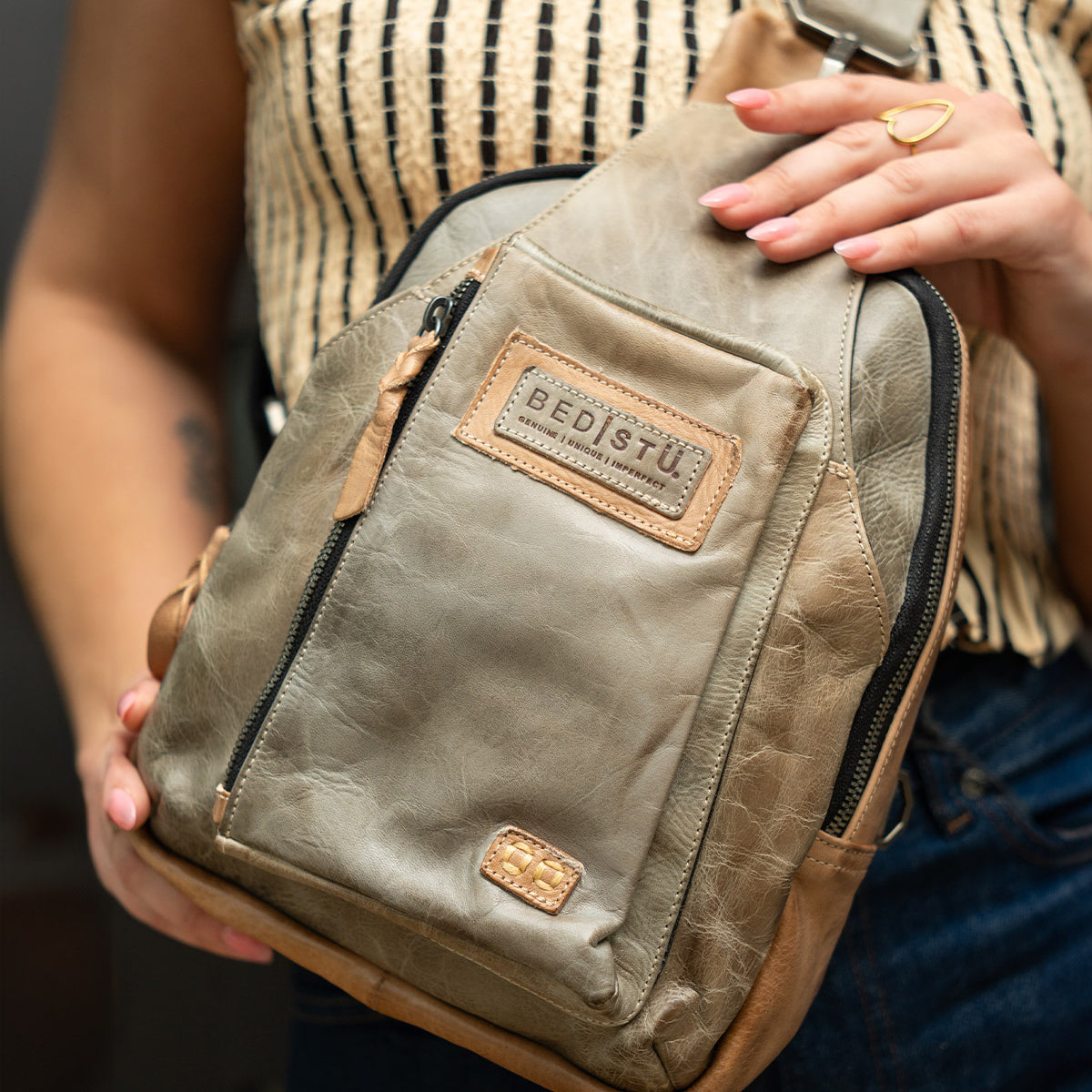 A person holds the Beau crossbody bag by Bed|Stü in tan leather—a versatile piece featuring a zip pocket that easily transforms into a compact backpack—styled with a patterned sleeveless top and dark jeans.