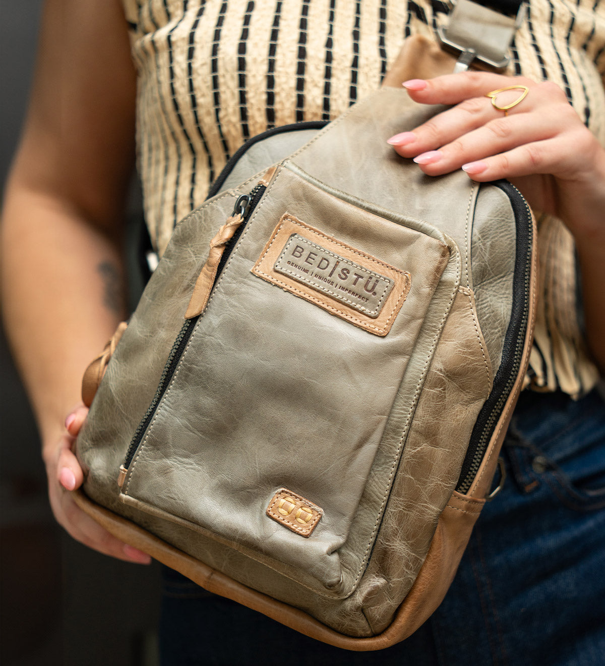A person holds the Beau crossbody bag by Bed|Stü in tan leather—a versatile piece featuring a zip pocket that easily transforms into a compact backpack—styled with a patterned sleeveless top and dark jeans.