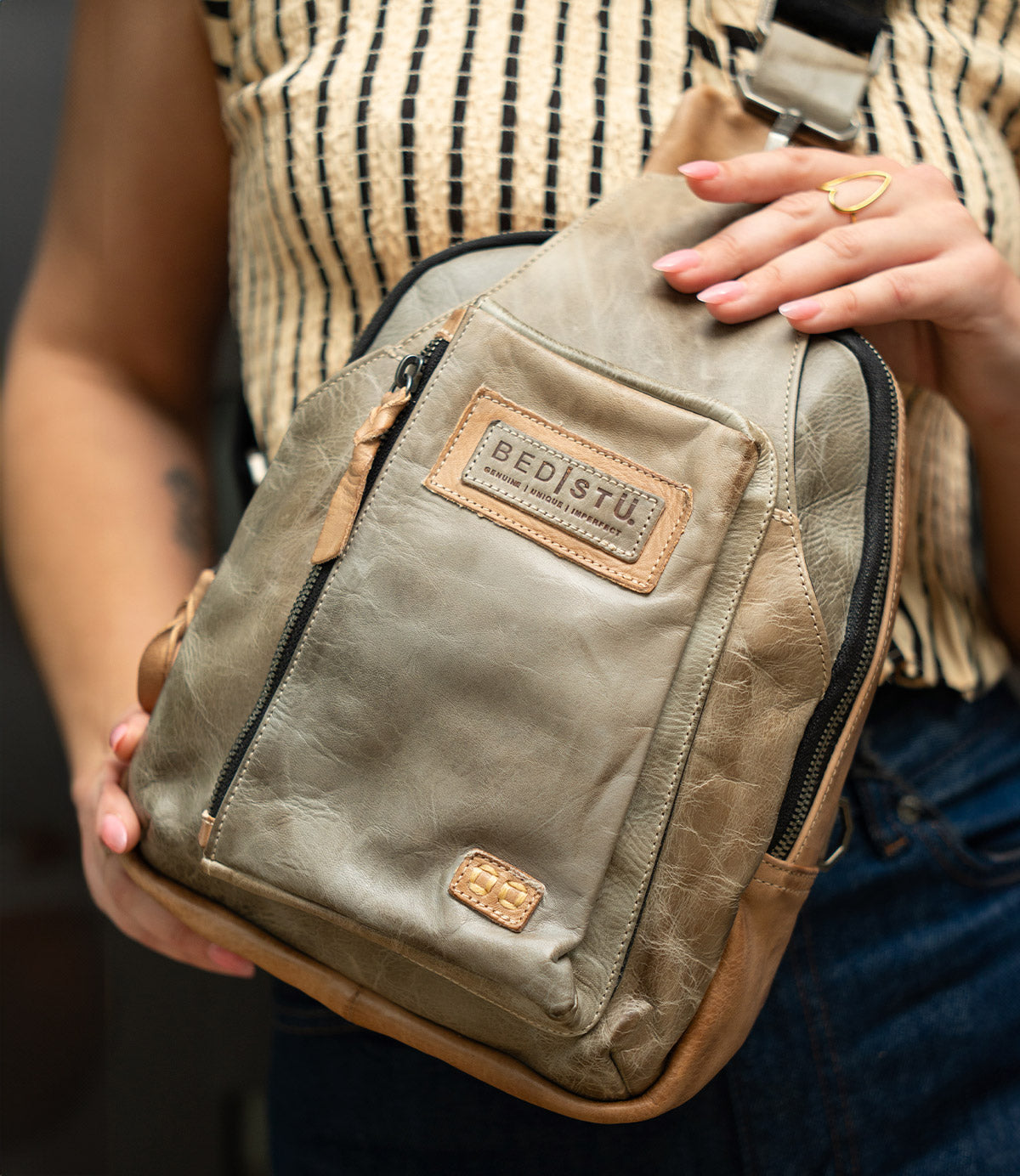 A person holds the Beau crossbody bag by Bed|Stü in tan leather—a versatile piece featuring a zip pocket that easily transforms into a compact backpack—styled with a patterned sleeveless top and dark jeans.