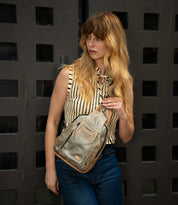 A woman with long, wavy hair stands in front of a dark paneled door, wearing a sleeveless striped top and jeans, carrying the Bed|Stü Beau sleek leather backpack over her shoulder.
