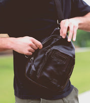 A person wearing a black shirt is opening the zipper of the sleek Bed|Stü Beau black leather sling backpack outdoors.