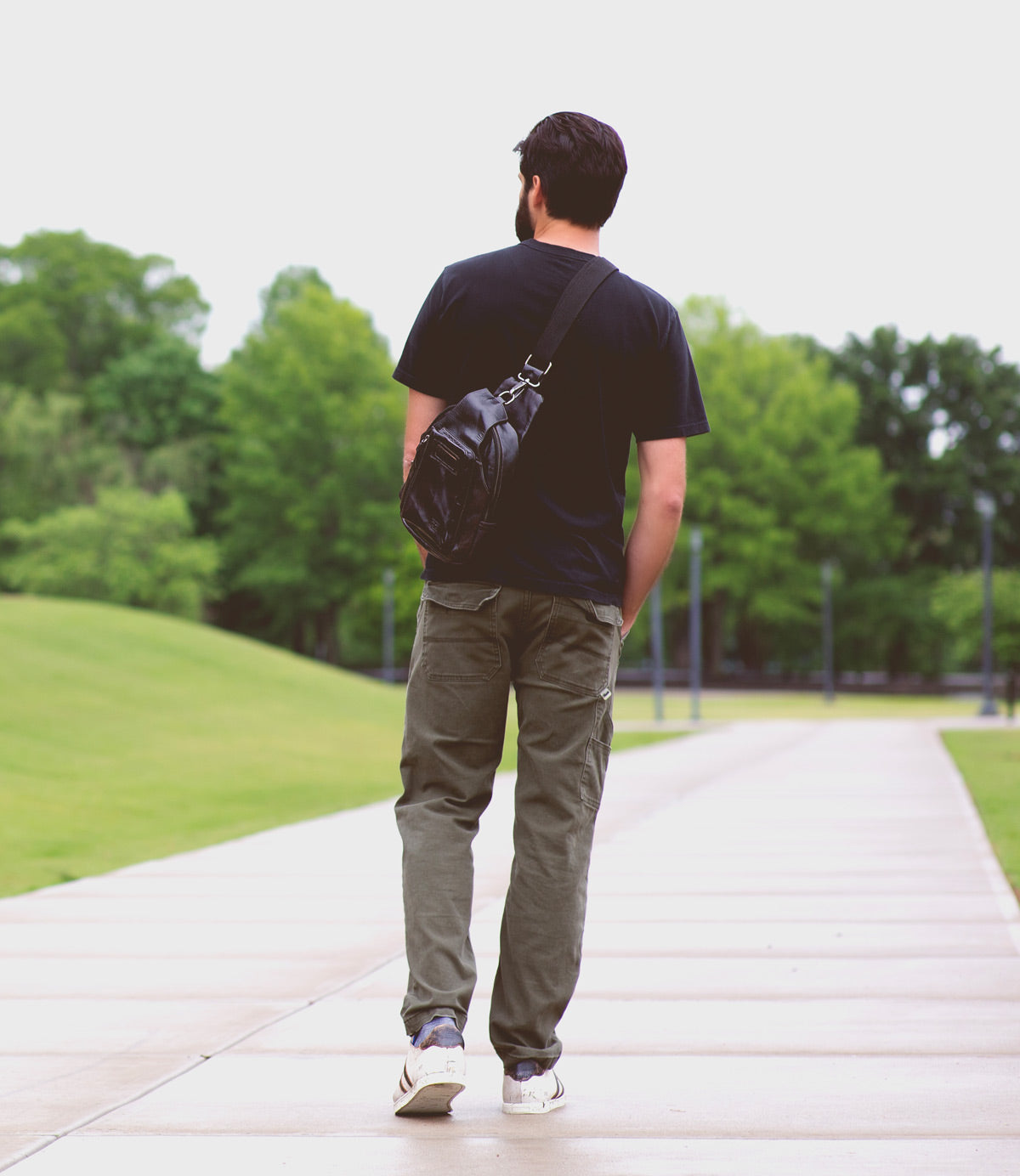 A man with the compact Beau backpack from Bed|Stü walks alone on a paved path through a green park, surrounded by trees and grass.