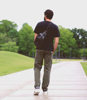 A man with the compact Beau backpack from Bed|Stü walks alone on a paved path through a green park, surrounded by trees and grass.