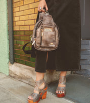 A person stands on a sidewalk in front of a brick wall, wearing Bed|Stü’s Beau gray platform sandals and a long black skirt, holding a small compact backpack.