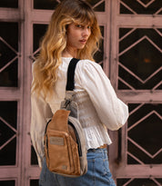 A woman wearing a white blouse and jeans stands outdoors with the Bed|Stü Beau brown and gray leather sling bag over her shoulder, facing slightly away from the camera.