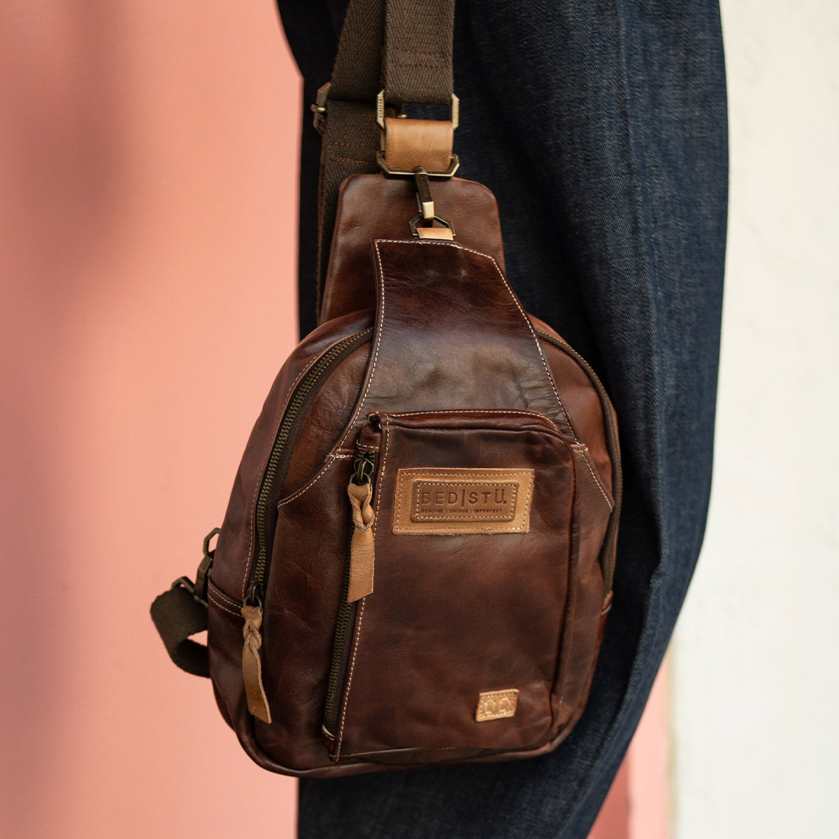 The Beau sling backpack by Bed|Stü, crafted in brown leather with a zipper pocket and a tan patch label, hangs on a person's shoulder against a pink and white background.