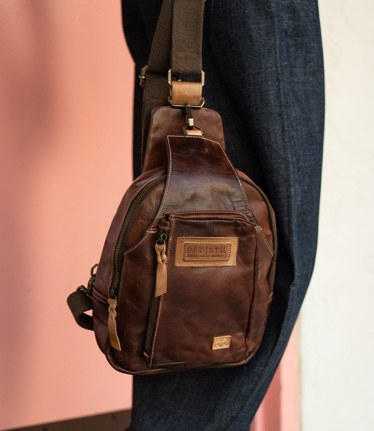 The Beau sling backpack by Bed|Stü, crafted in brown leather with a zipper pocket and a tan patch label, hangs on a person's shoulder against a pink and white background.