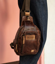 The Beau sling backpack by Bed|Stü, crafted in brown leather with a zipper pocket and a tan patch label, hangs on a person's shoulder against a pink and white background.