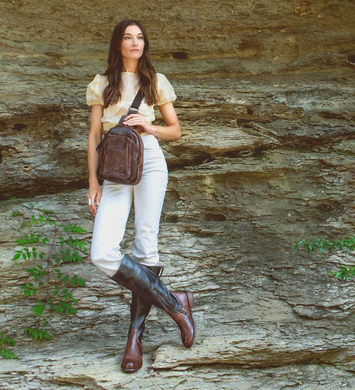 A woman stands against a rocky wall, wearing a light blouse, white pants, knee-high brown boots, and carrying the stylish Bed|Stü Beau leather sling bag. Green plants grow at the base of the rock.