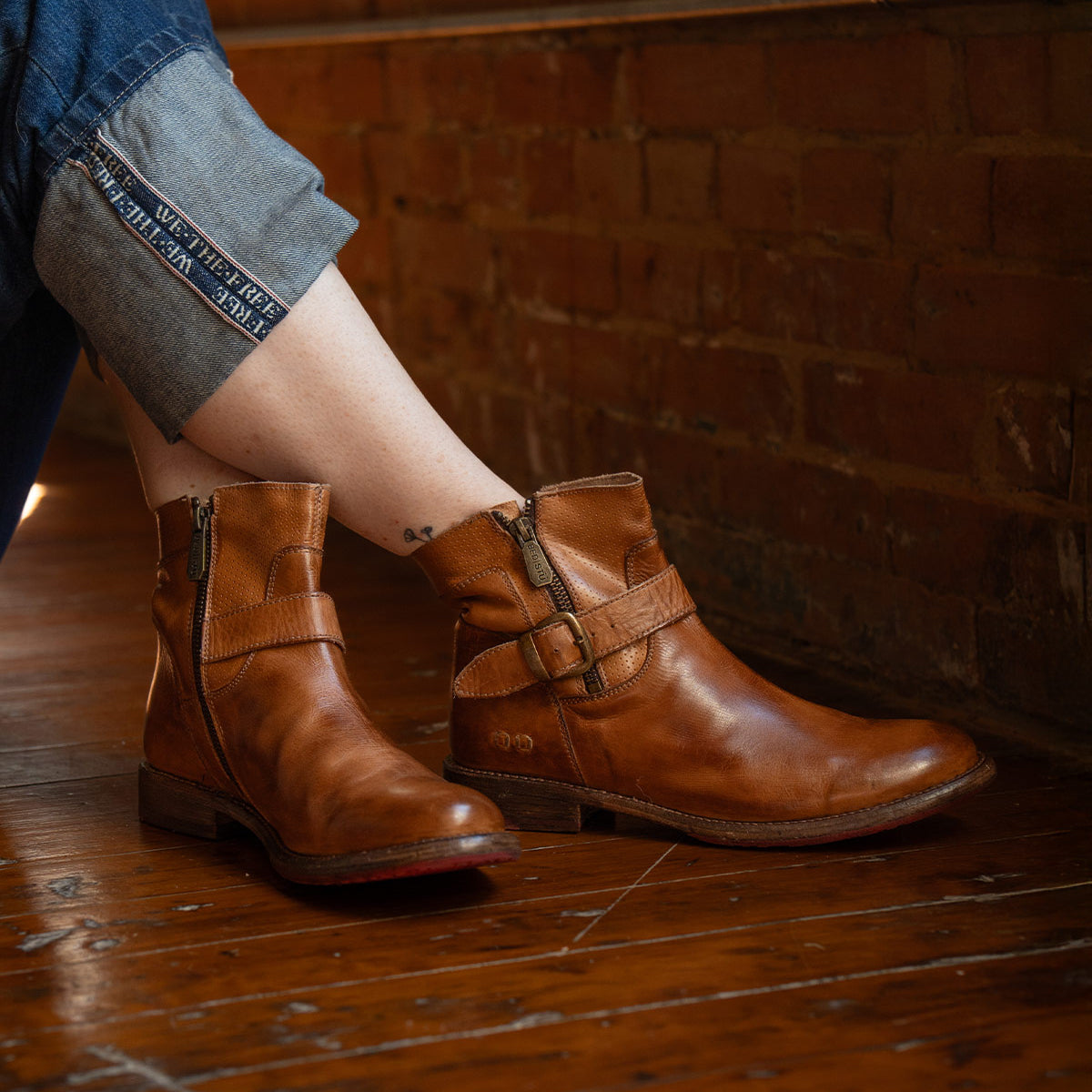 Wearing rolled-up jeans, the person sits on a wooden floor against a brick wall, showing off Bed|Stü’s Becca brown leather ankle boots with a sturdy leather shaft and an ankle buckle.