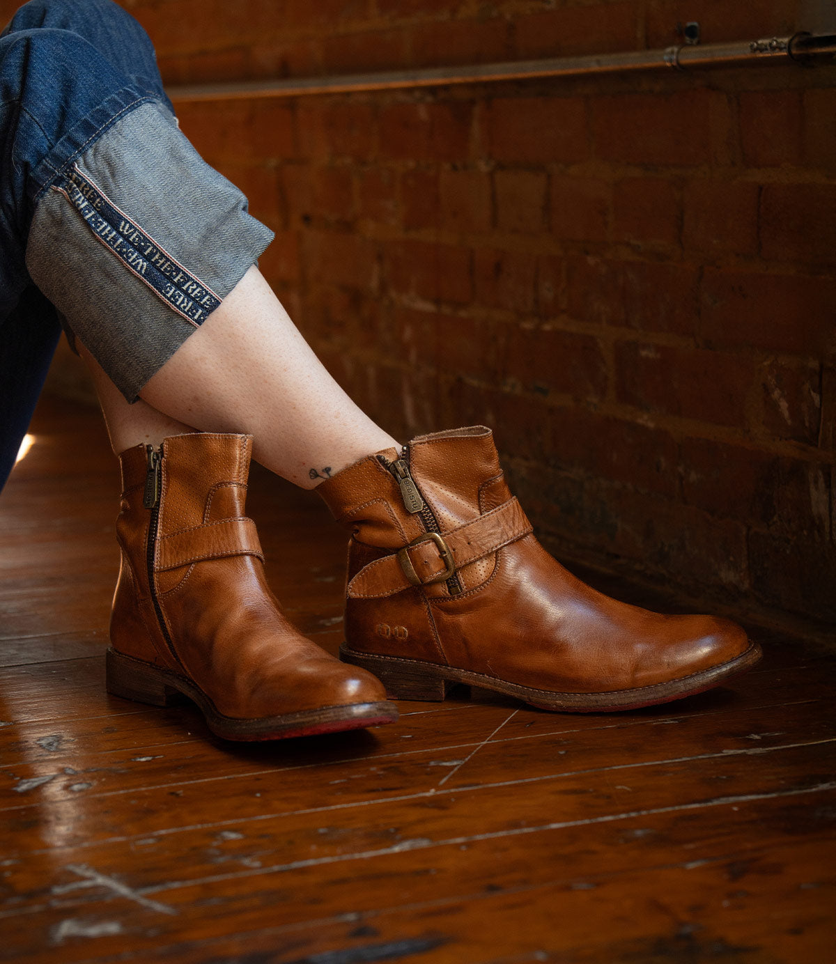 Wearing rolled-up jeans, the person sits on a wooden floor against a brick wall, showing off Bed|Stü’s Becca brown leather ankle boots with a sturdy leather shaft and an ankle buckle.