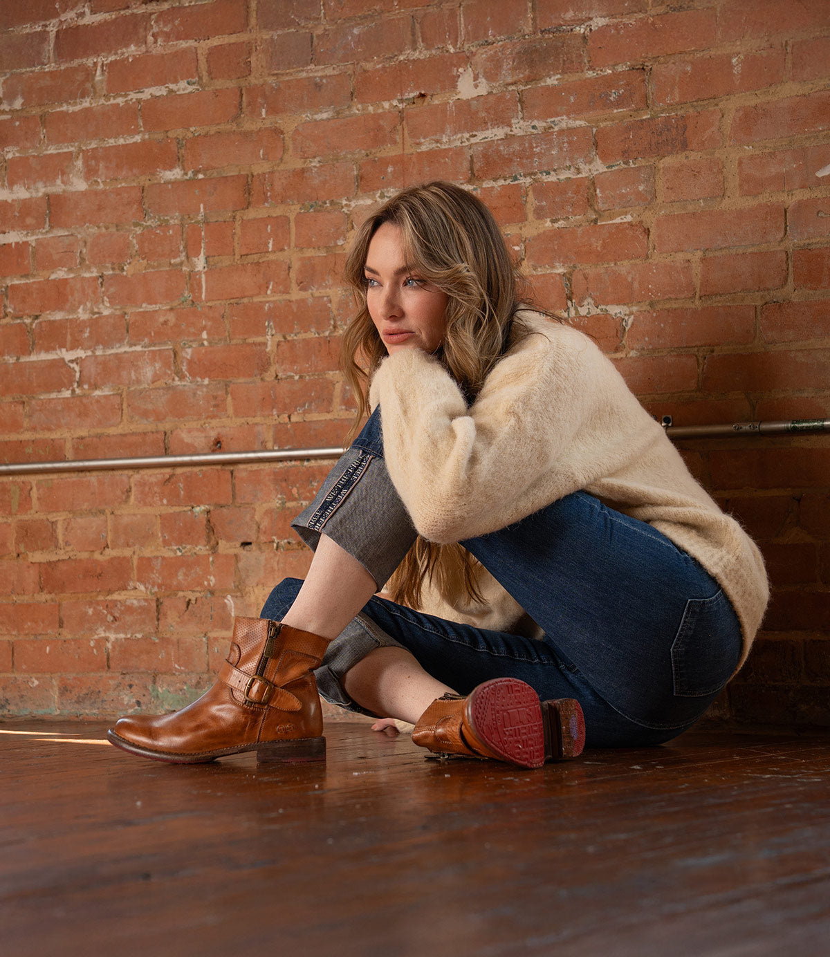 A woman in a beige sweater and jeans sits on a wooden floor against a brick wall, resting her chin on her hand and gazing to the side, her feet clad in Bed|Stü's Becca boots, featuring sturdy leather shafts and ankle buckles.