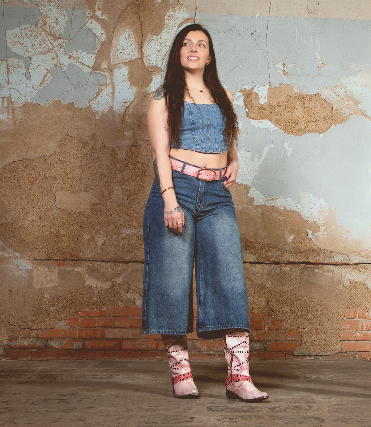 A young woman with long dark hair stands in front of a distressed wall. She wears a denim crop top, wide-leg jeans, and the Bed|Stü Betting handcrafted leather boots featuring subtle floral inlays.