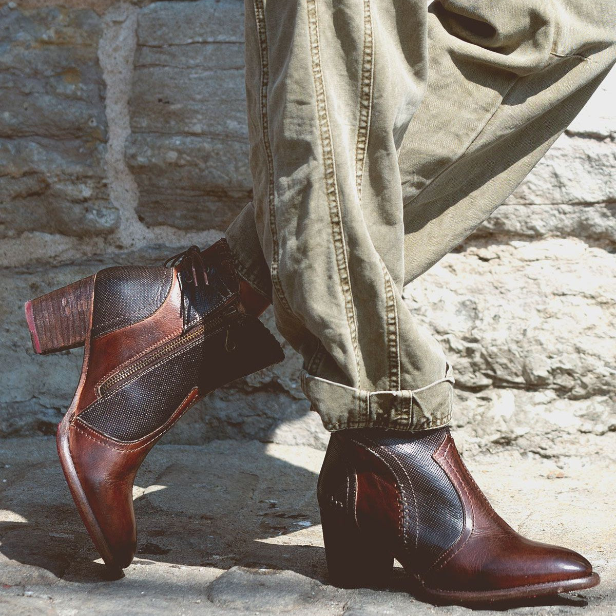 A person wearing cuffed khaki pants and the Bed|Stü Bia two-tone brown handcrafted leather ankle boots stands on cobblestone pavement in front of a stone wall.