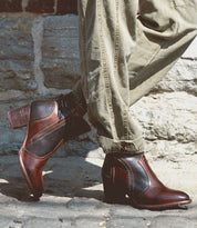 A person wearing cuffed khaki pants and the Bed|Stü Bia two-tone brown handcrafted leather ankle boots stands on cobblestone pavement in front of a stone wall.