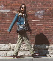 A woman wearing sunglasses, a denim jacket, khaki pants, and the handcrafted Bed|Stü Bia leather ankle boots walks on a sidewalk beside a brick wall, carrying a black shoulder bag.