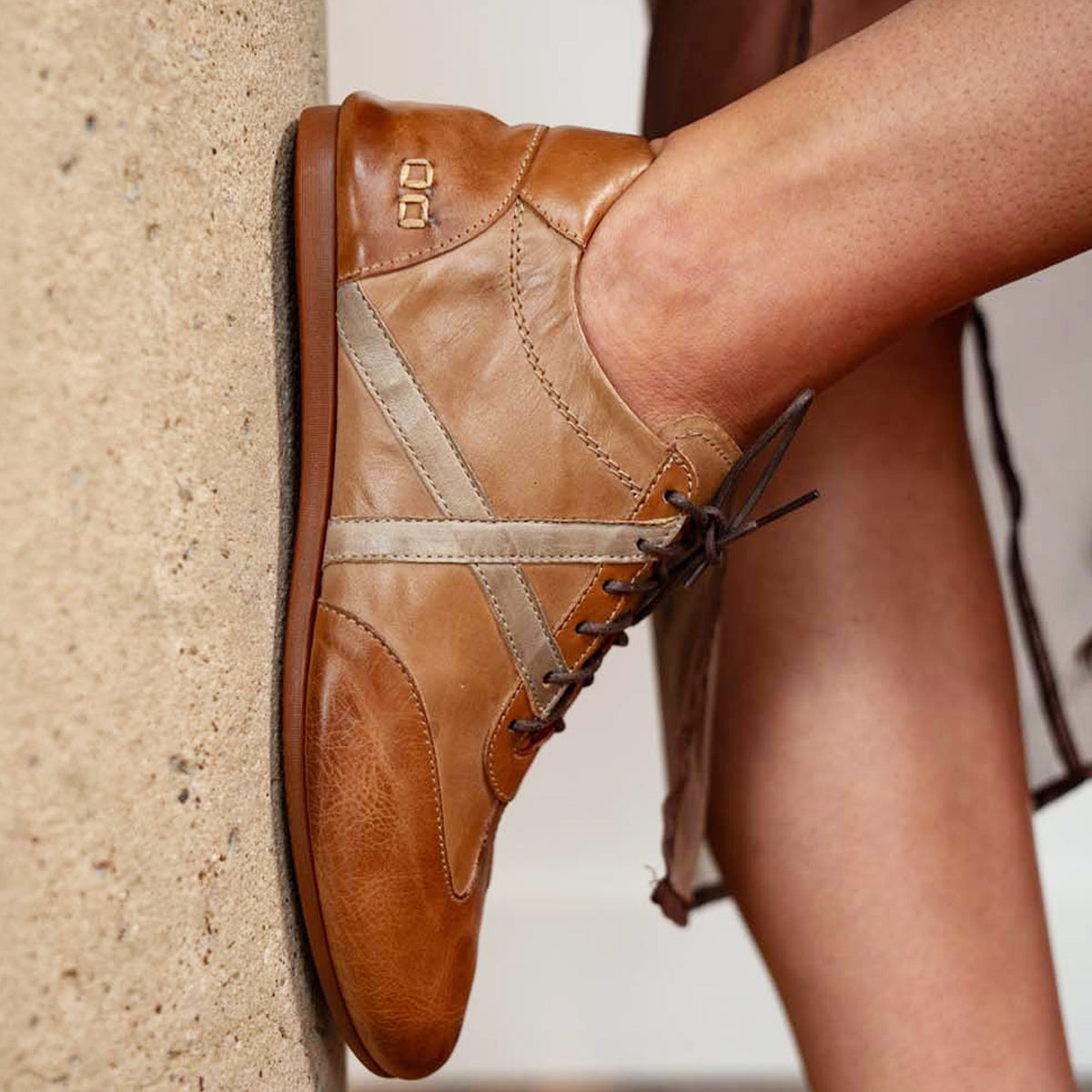Close-up of a person wearing the Bed|Stü Bingo II sneaker in tan and beige leather with laces, featuring an XL EXTRALIGHT outsole, resting their foot against a textured concrete surface.