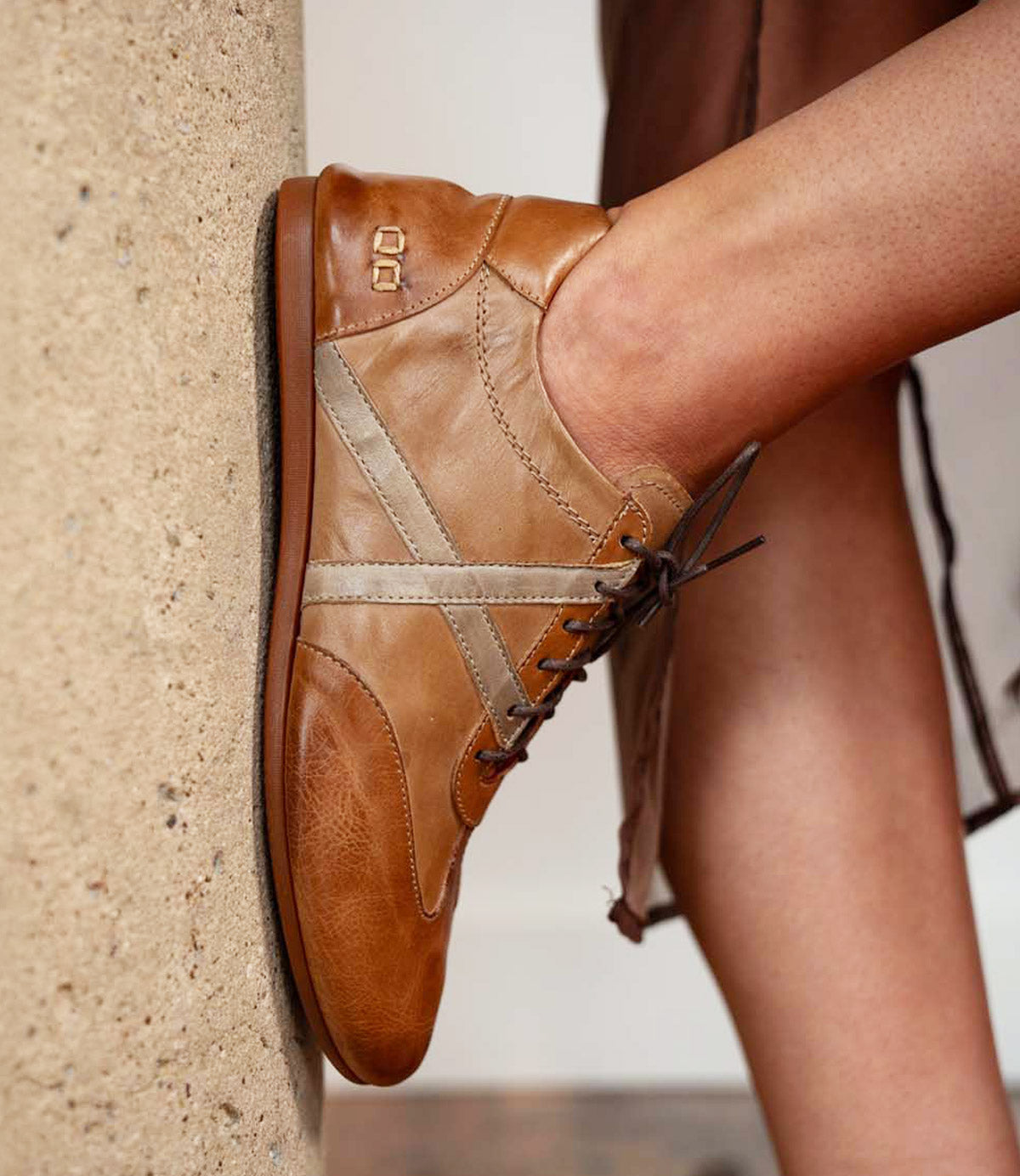 Close-up of a person wearing the Bed|Stü Bingo II sneaker in tan and beige leather with laces, featuring an XL EXTRALIGHT outsole, resting their foot against a textured concrete surface.