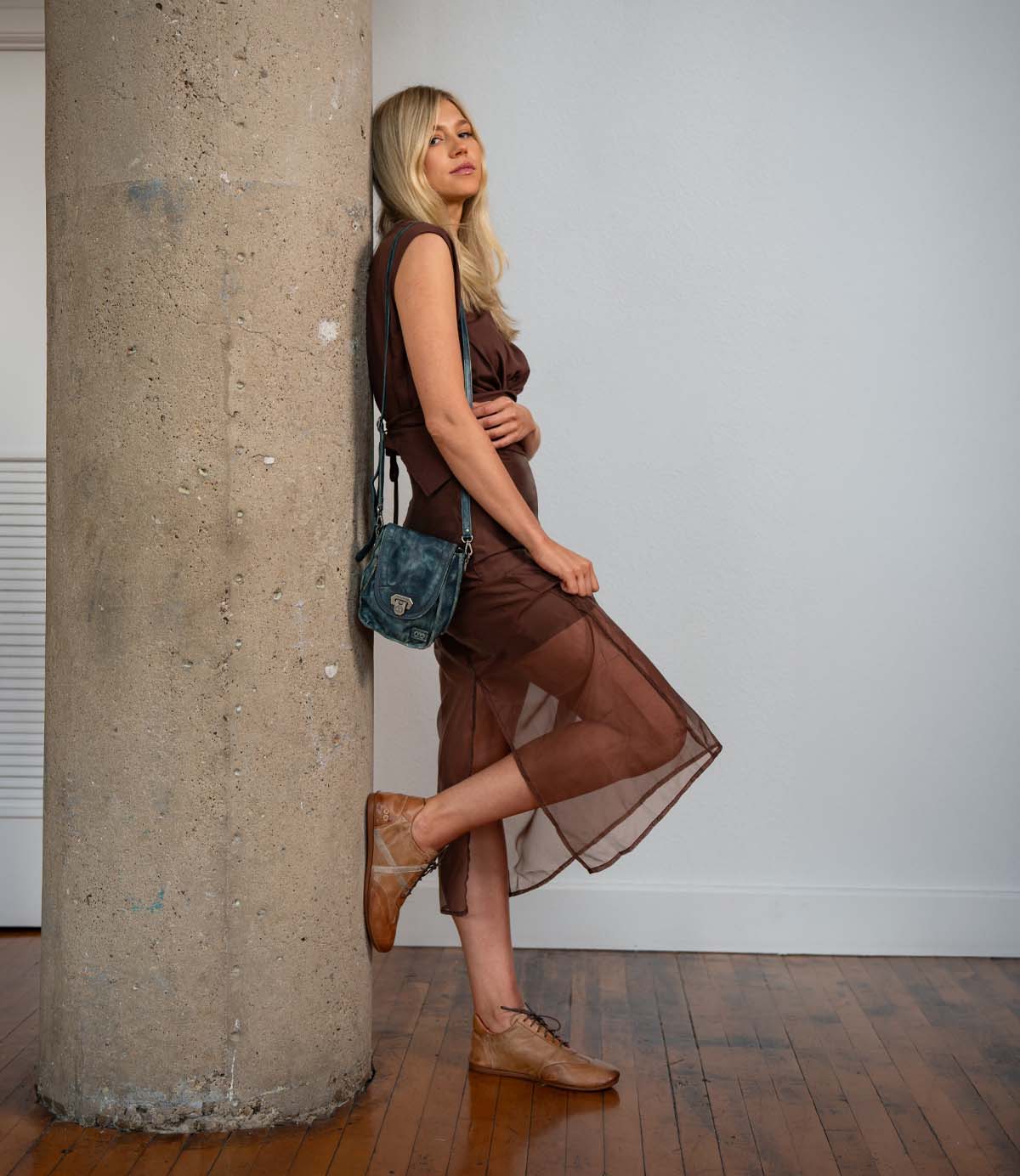 A woman in a sheer brown dress and brown shoes—the Bingo II by Bed|Stü, featuring an EXTRALIGHT outsole—leans against a large concrete pillar indoors, holding a dark satchel bag and standing on a wooden floor.