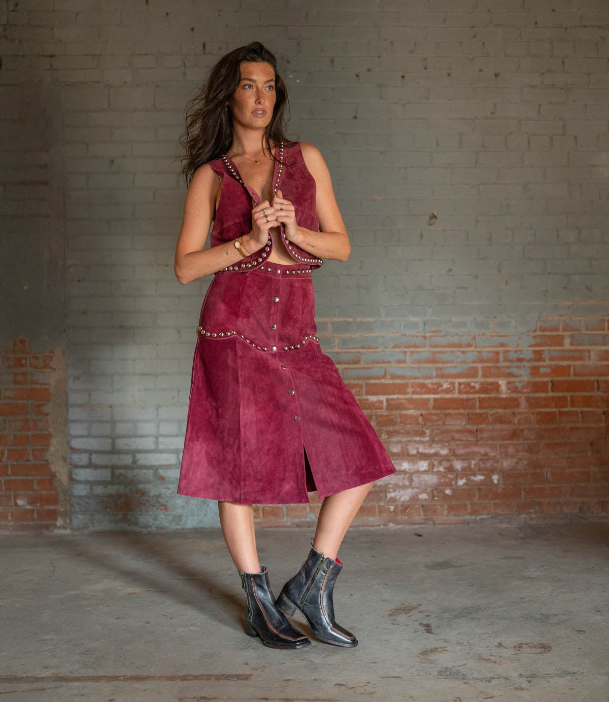 A woman stands indoors against a brick wall, wearing a sleeveless maroon dress with metal studs and Bed|Stü Bitsy black leather boots featuring a square toe and stacked heel.
