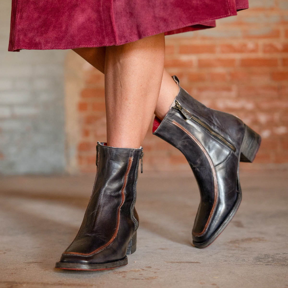 A person stands on a concrete floor wearing the Bed|Stü Bitsy black leather ankle boots, featuring a stacked heel and side zippers, paired with a burgundy skirt.