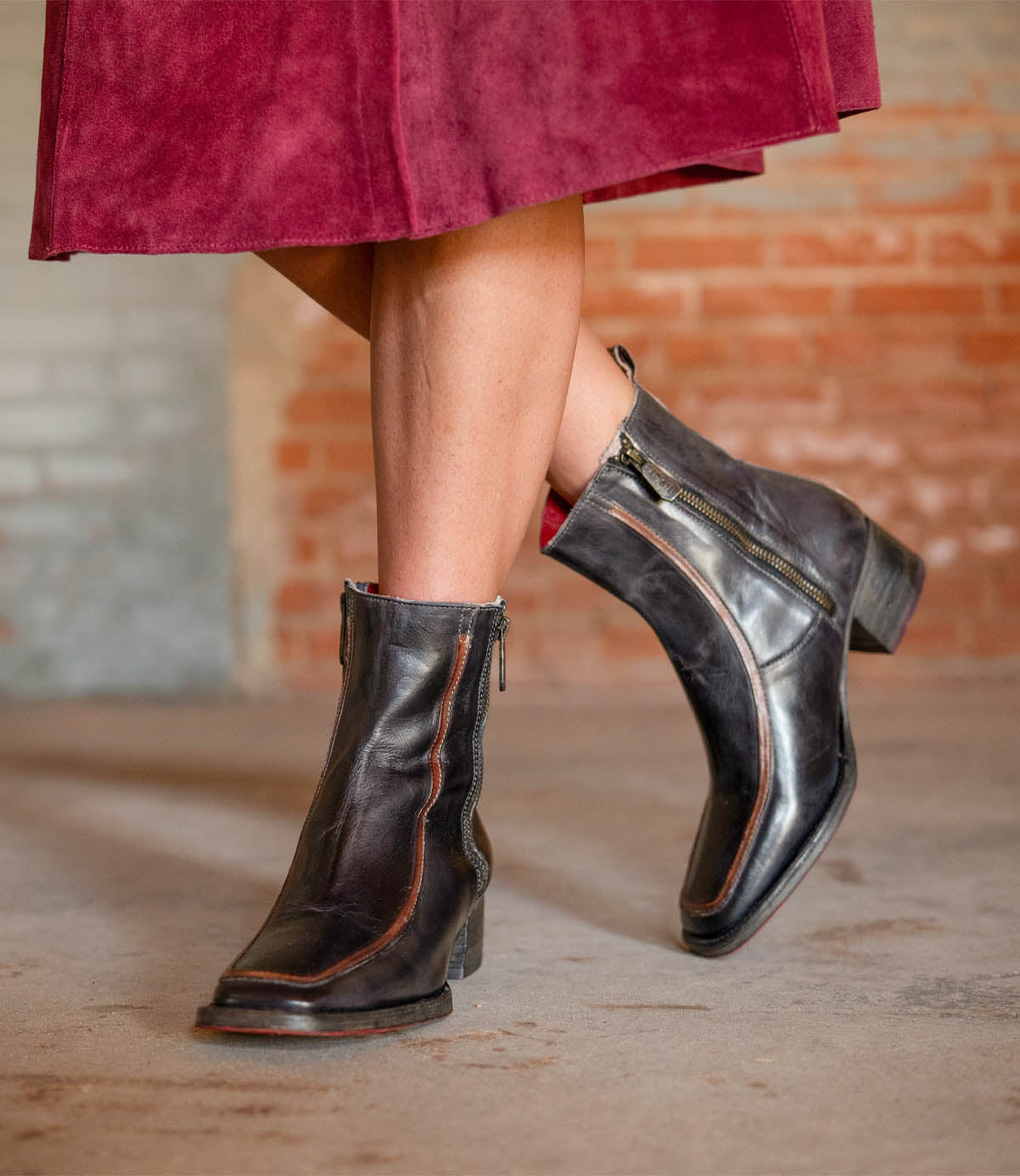 A person stands on a concrete floor wearing the Bed|Stü Bitsy black leather ankle boots, featuring a stacked heel and side zippers, paired with a burgundy skirt.